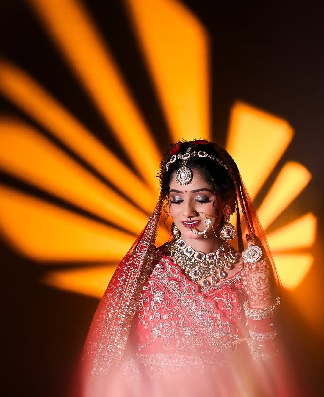 Indian bride in red attire and jewelry posing against radiating orange light beams.