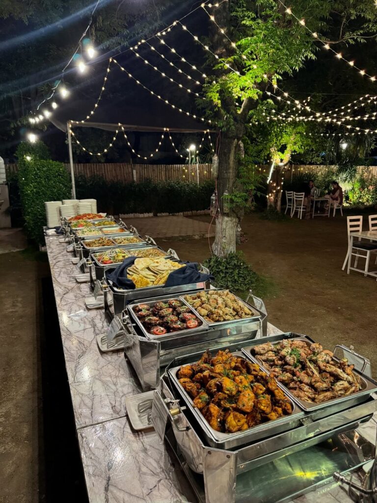Buffet table with silver chafing dishes of food under outdoor string lights