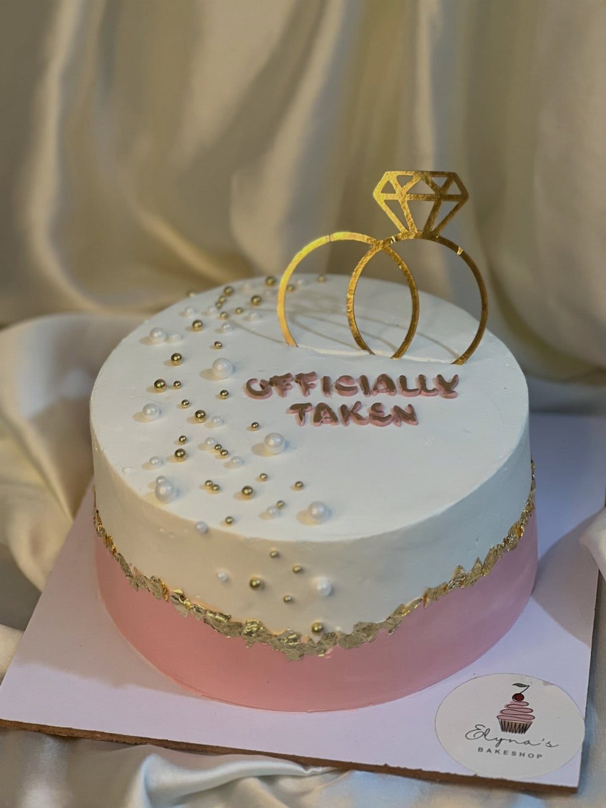 Engagement cake with white and pink frosting, gold ring topper, and 'Officially Taken' text.