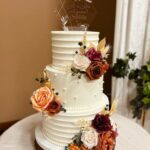 Three-tier white wedding cake decorated with autumn-colored roses on a rustic wood slice.