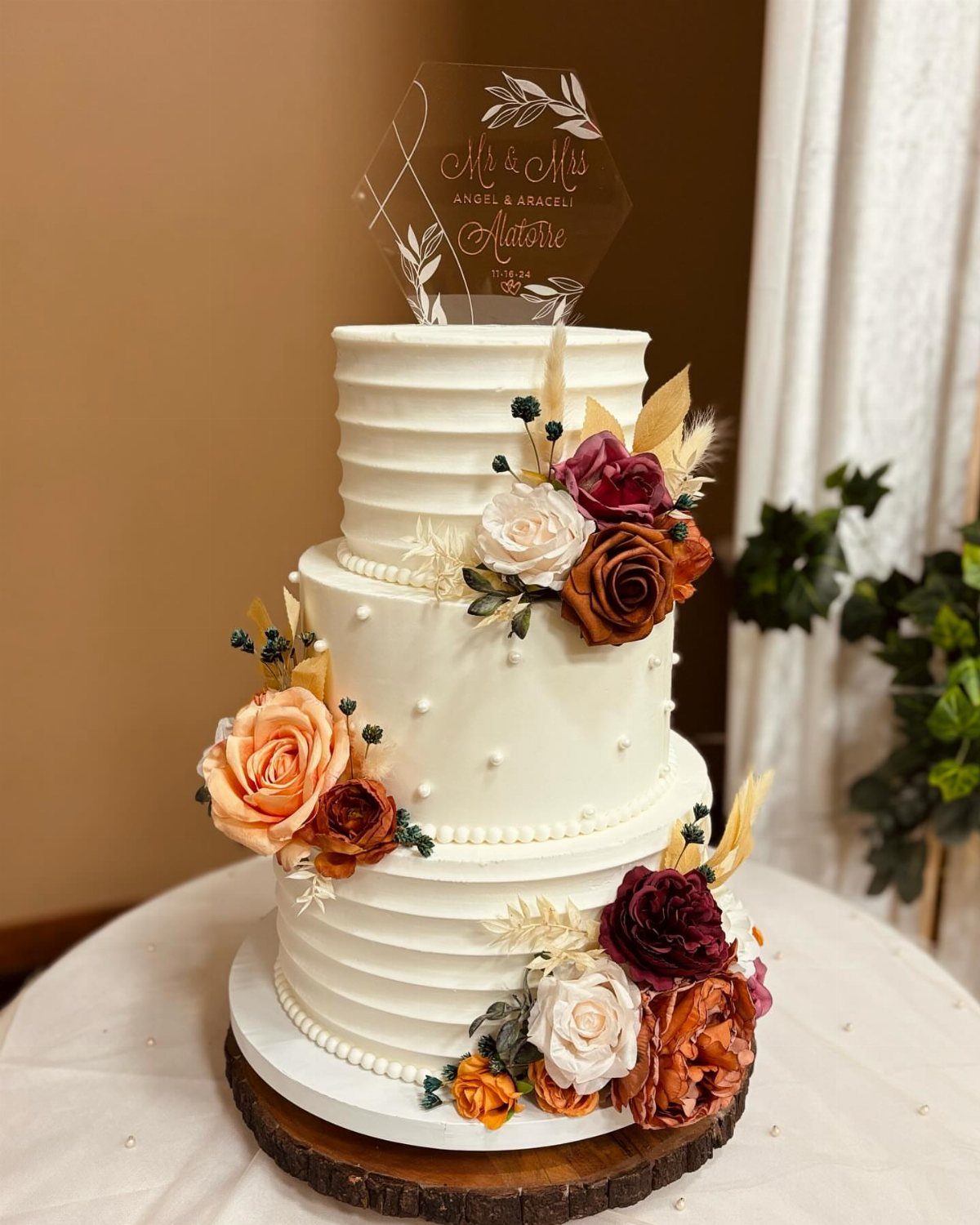 Three-tier white wedding cake decorated with autumn-colored roses on a rustic wood slice.