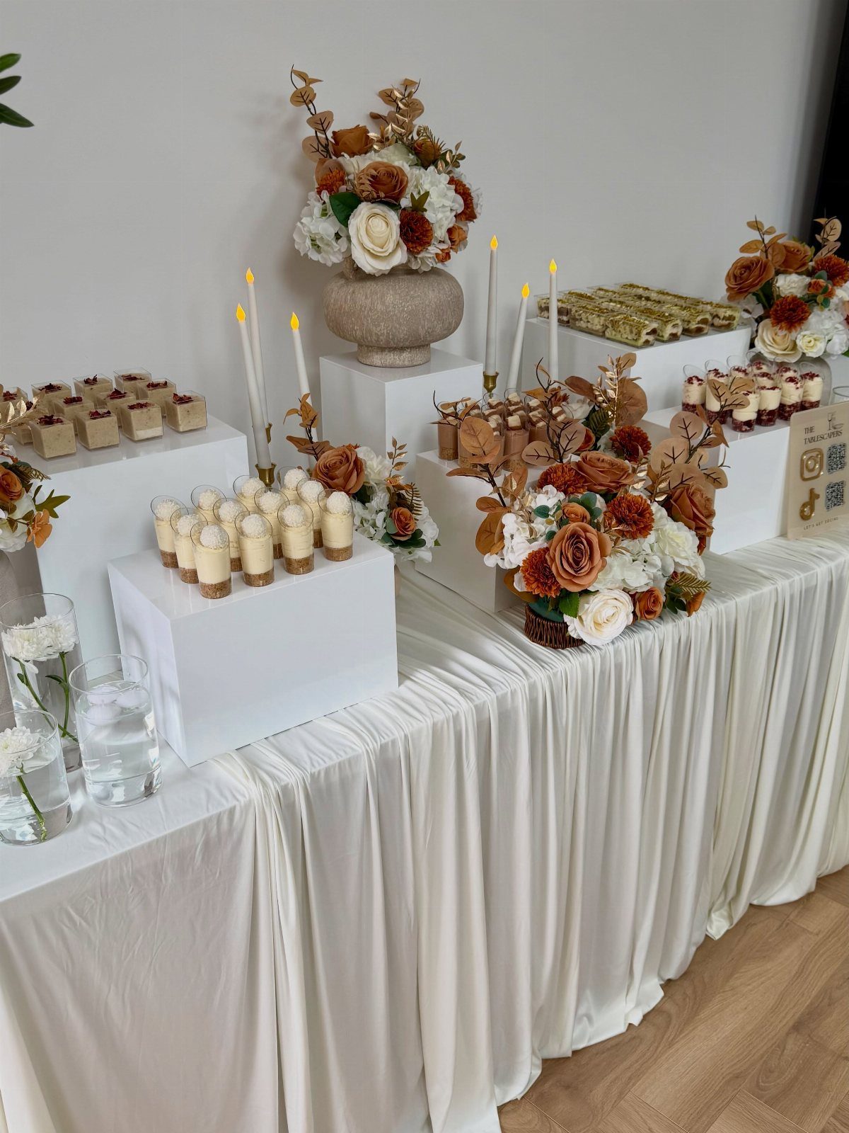 Dessert table with autumn floral arrangements and small cakes on white pedestals.