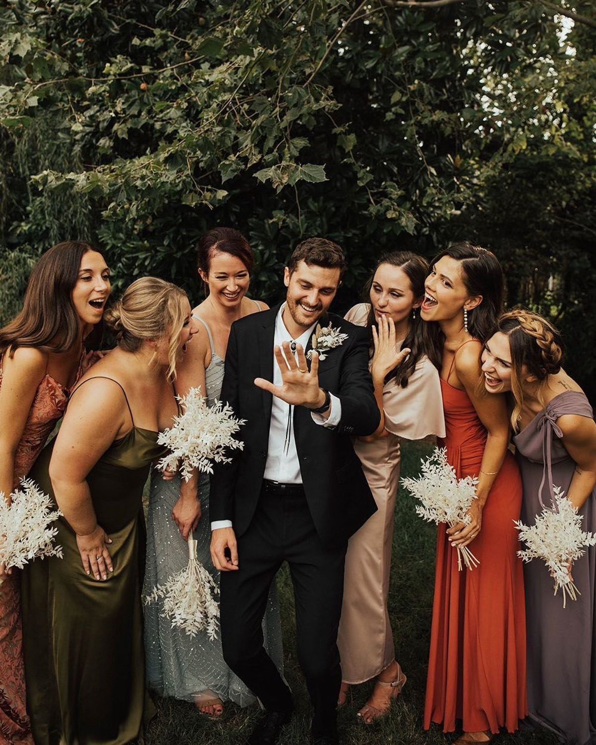 Groom showing his wedding ring to laughing bridesmaids in a lush green garden.
