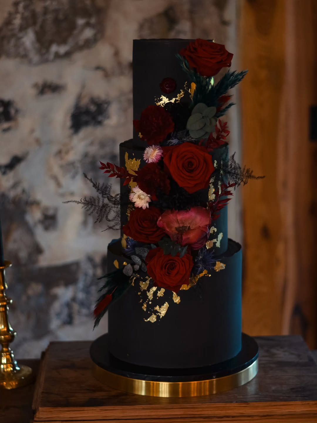 Three-tier matte black cake adorned with red roses and gold leaf accents.