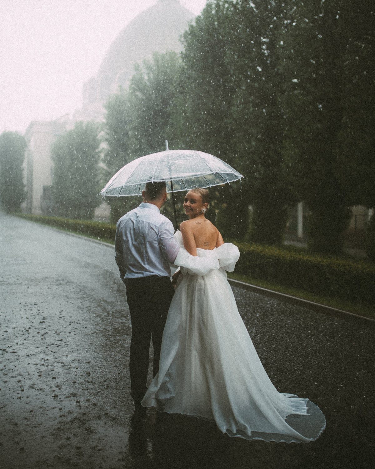 Bride and groom standing under a clear umbrella on a rainy road.