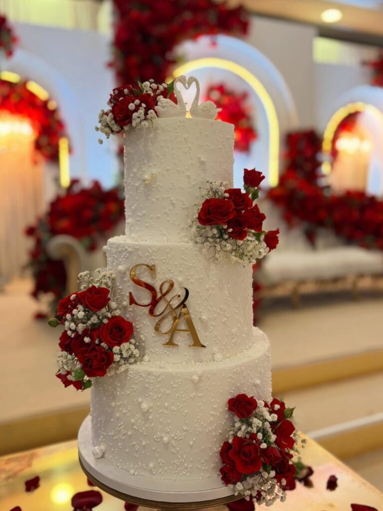 Three-tiered white wedding cake decorated with red roses and gold initials on a stand.