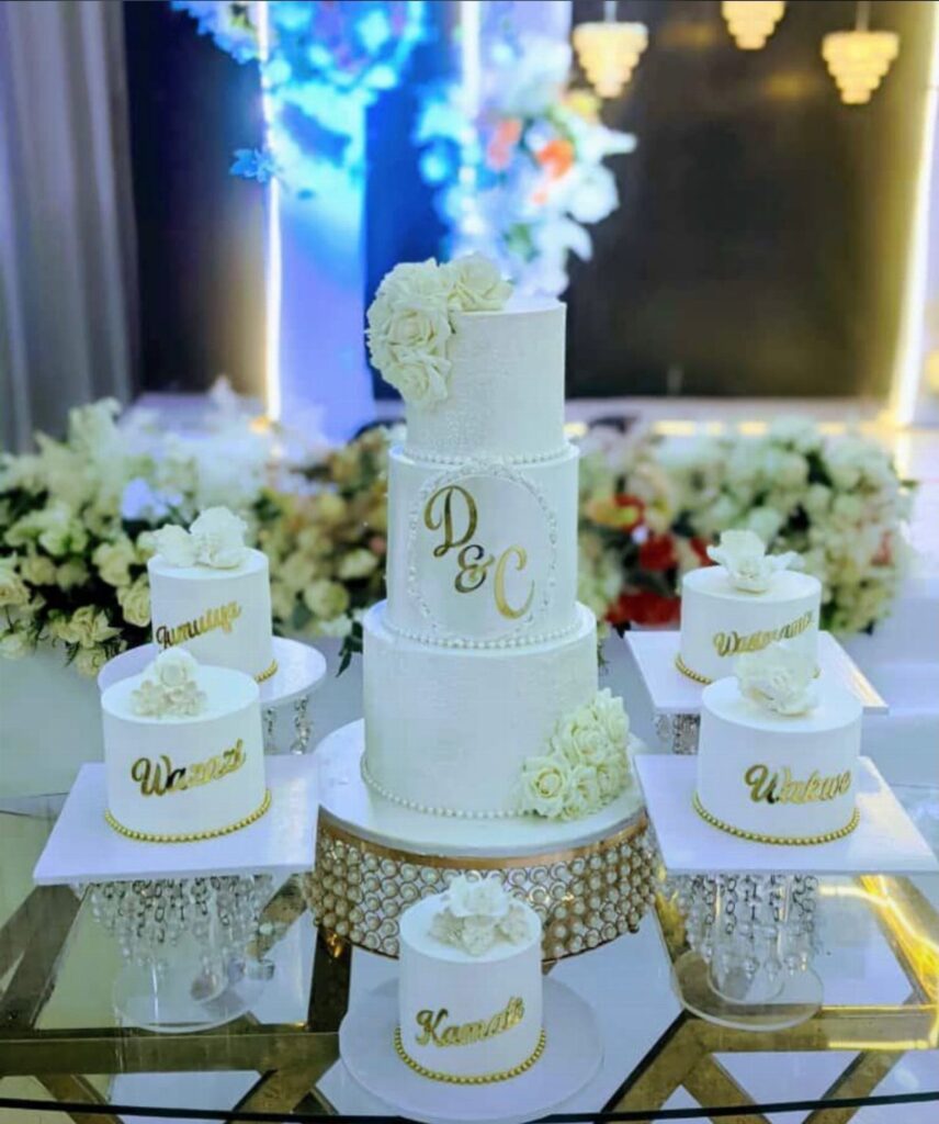 Three-tier white wedding cake with gold initials and surrounding smaller white cakes.