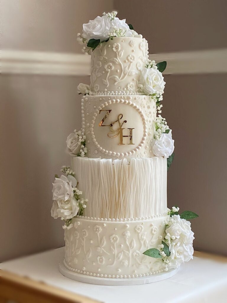Four-tier white wedding cake with intricate frosting, white roses, and gold monogram initials.