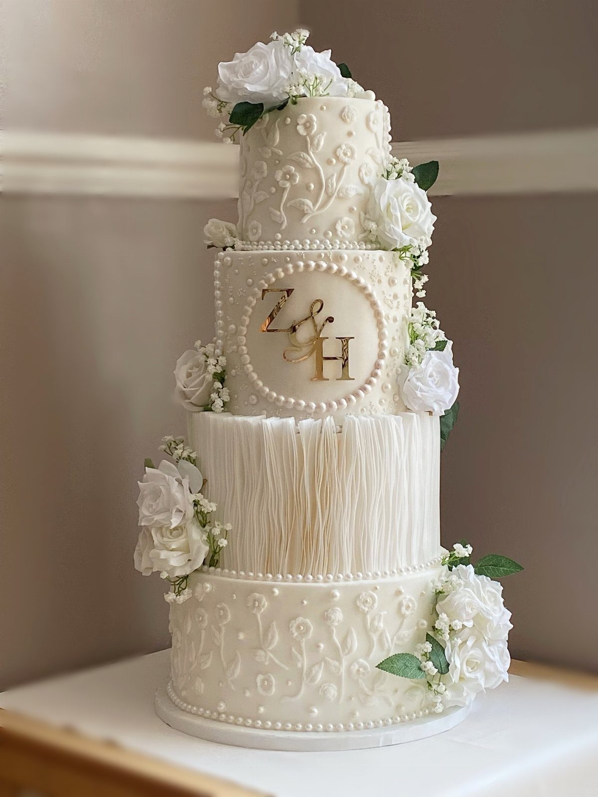 Four-tier white wedding cake with intricate frosting, white roses, and gold monogram initials.