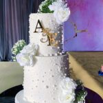 Two-tier white wedding cake with pearl decorations, white roses, and gold initials.
