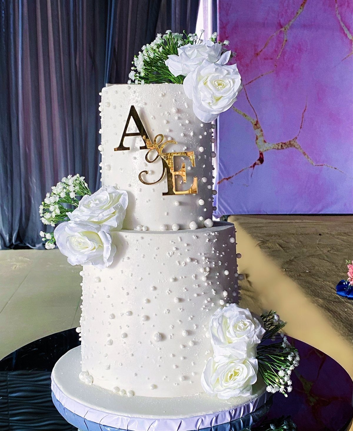 Two-tier white wedding cake with pearl decorations, white roses, and gold initials.