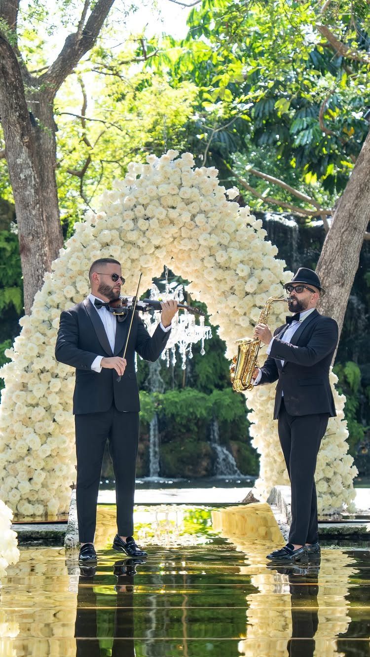 Musicians playing violin and saxophone in front of a white floral wedding arch.