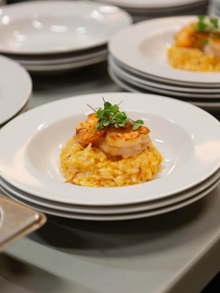 Shrimp and yellow risotto served on a white plate among stacked dishes.