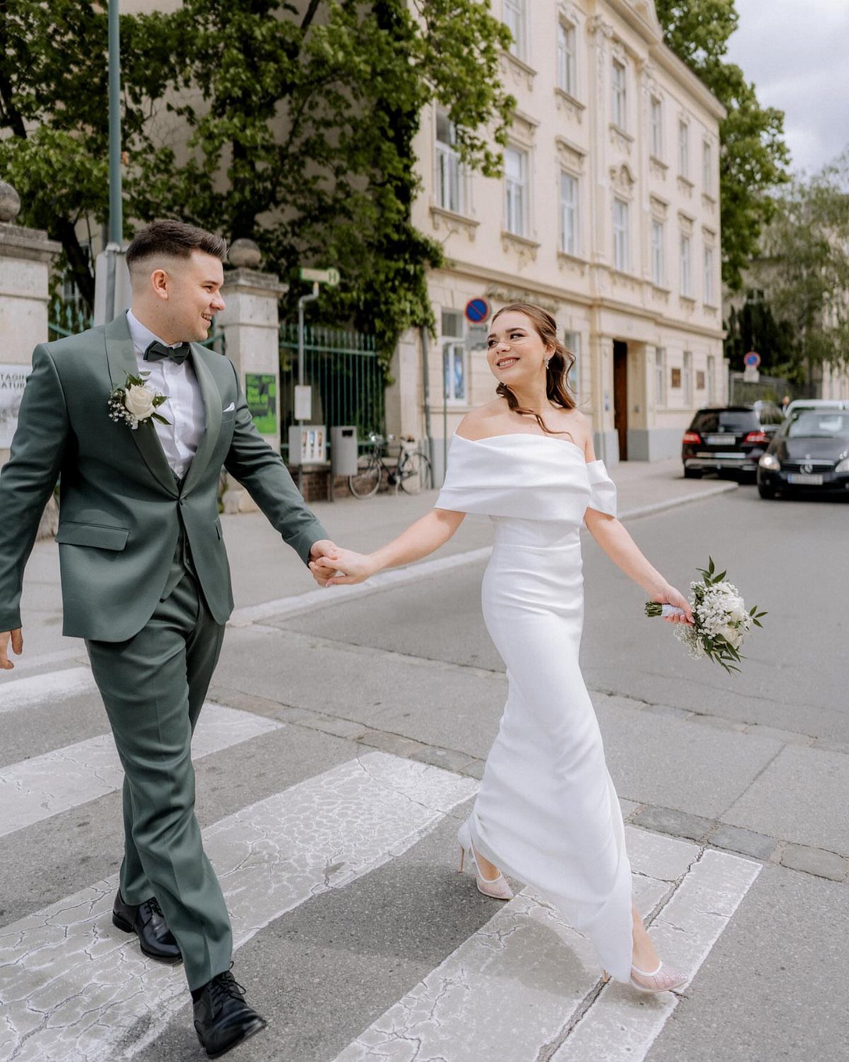 Bride and groom holding hands while crossing a city street in wedding attire.