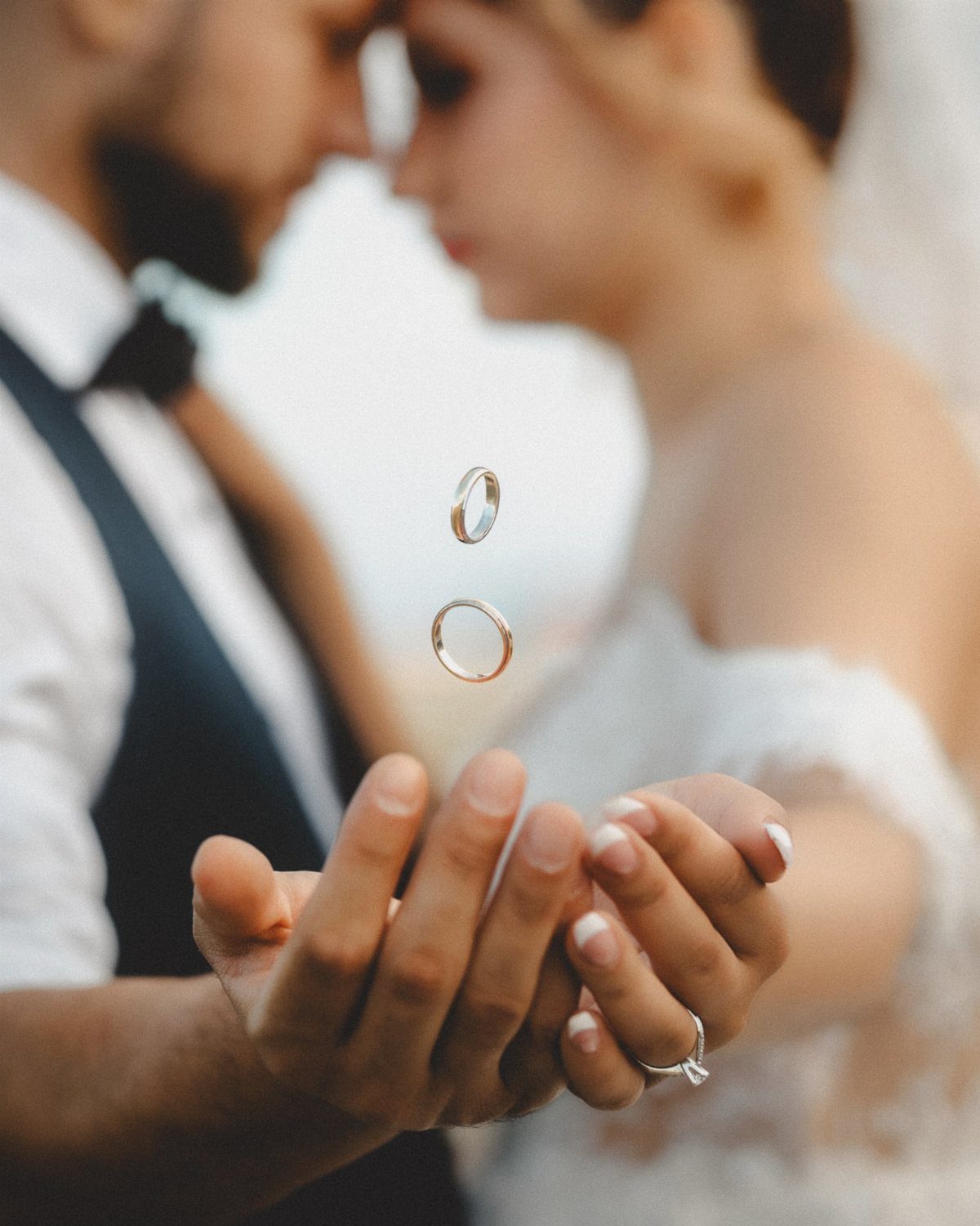 Two gold wedding rings floating above the hands of a blurred bride and groom.